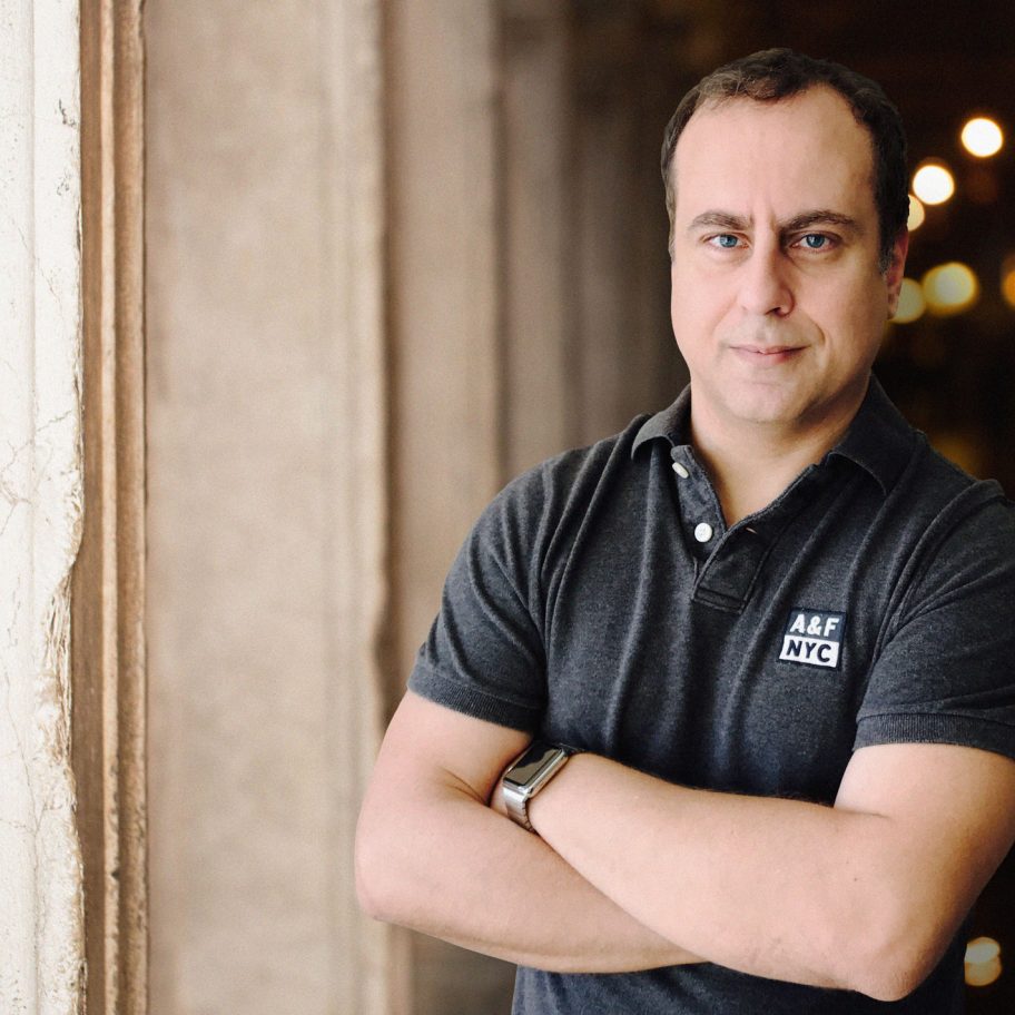 A man in a dark gray polo shirt with folded arms is leaning against a light-colored stone wall. He has short hair and is looking directly at the camera with a slight smile. The background is blurred, featuring a corridor with glowing lights.