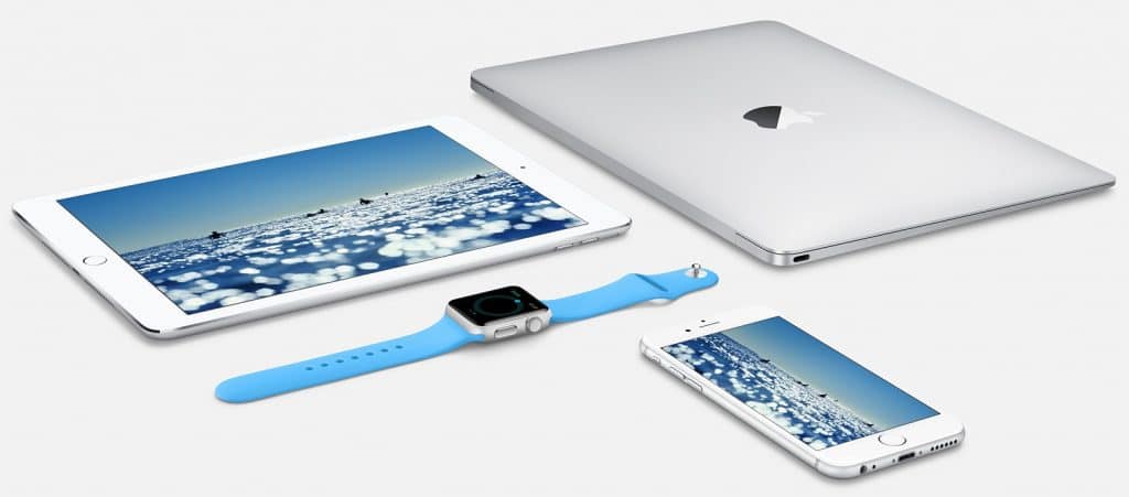 An assortment of Apple devices on a white surface including a silver MacBook, a silver iPad displaying a blue water scene, a silver iPhone showing the same water scene, and an Apple Watch with a blue band displaying a black screen, showcasing the seamless integration of the Apple ecosystem.