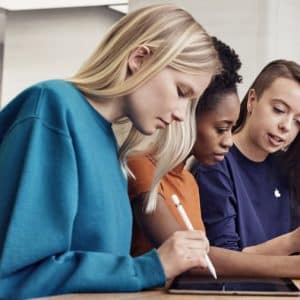 Three individuals are seated at a table, using Apple tablets with styluses. The person on the left has long blonde hair and is wearing a blue top. The middle person has curly dark hair and is wearing an orange top. The person on the right has long, partially shaved hair and is wearing a navy top.