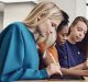 Three individuals are seated at a table, using Apple tablets with styluses. The person on the left has long blonde hair and is wearing a blue top. The middle person has curly dark hair and is wearing an orange top. The person on the right has long, partially shaved hair and is wearing a navy top.