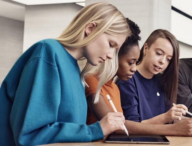 Three individuals are seated at a table, using Apple tablets with styluses. The person on the left has long blonde hair and is wearing a blue top. The middle person has curly dark hair and is wearing an orange top. The person on the right has long, partially shaved hair and is wearing a navy top.