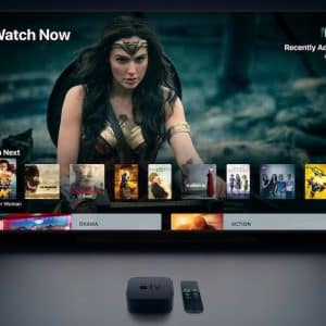 A television screen displays a streaming service interface with a selection of movies, highlighting Apple's new original programming launched in March 2019. A scene from "Wonder Woman" is prominently featured at the top. Below, categories like "Up Next" and genres such as "Drama" and "Action" show several movie thumbnails. An Apple TV device and remote are visible on a table below the screen.