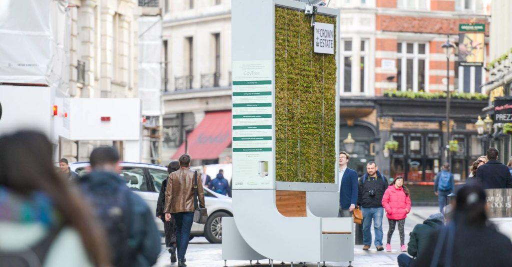 A bustling city street with people walking by a large, vertical urban installation covered in greenery. The installation includes an information panel on the side and is surrounded by historic buildings. Passersby are stopping to look and take photos, showcasing London’s fight against air pollution.