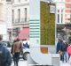 A bustling city street with people walking by a large, vertical urban installation covered in greenery. The installation includes an information panel on the side and is surrounded by historic buildings. Passersby are stopping to look and take photos, showcasing London’s fight against air pollution.