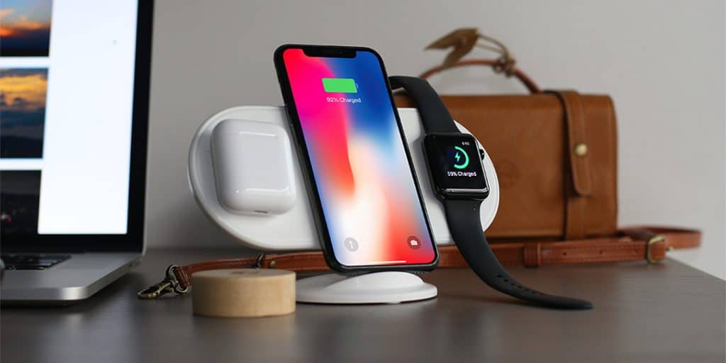 A smartphone, smartwatch, and wireless earbuds charging on a white wireless charging station, reminiscent of the innovative AirPower, on a desk. The phone, displaying a 90% charge, stands vertical. A leather bag and a laptop are in the background. Available end of March.