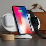 A smartphone, smartwatch, and wireless earbuds charging on a white wireless charging station, reminiscent of the innovative AirPower, on a desk. The phone, displaying a 90% charge, stands vertical. A leather bag and a laptop are in the background. Available end of March.