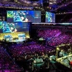 A large crowd in China watches an intense esports tournament in an arena. Multiple large screens display the game in progress, where several female professional gamers are competing. The stage is set up with gaming stations and illuminated with colorful lighting, creating an energetic atmosphere.