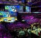 A large crowd in China watches an intense esports tournament in an arena. Multiple large screens display the game in progress, where several female professional gamers are competing. The stage is set up with gaming stations and illuminated with colorful lighting, creating an energetic atmosphere.