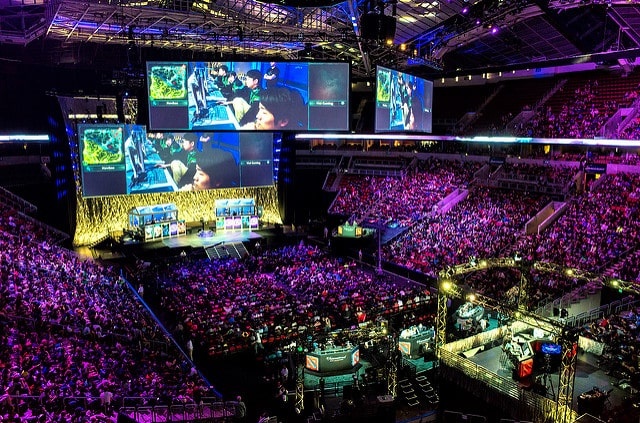 A large crowd in China watches an intense esports tournament in an arena. Multiple large screens display the game in progress, where several female professional gamers are competing. The stage is set up with gaming stations and illuminated with colorful lighting, creating an energetic atmosphere.