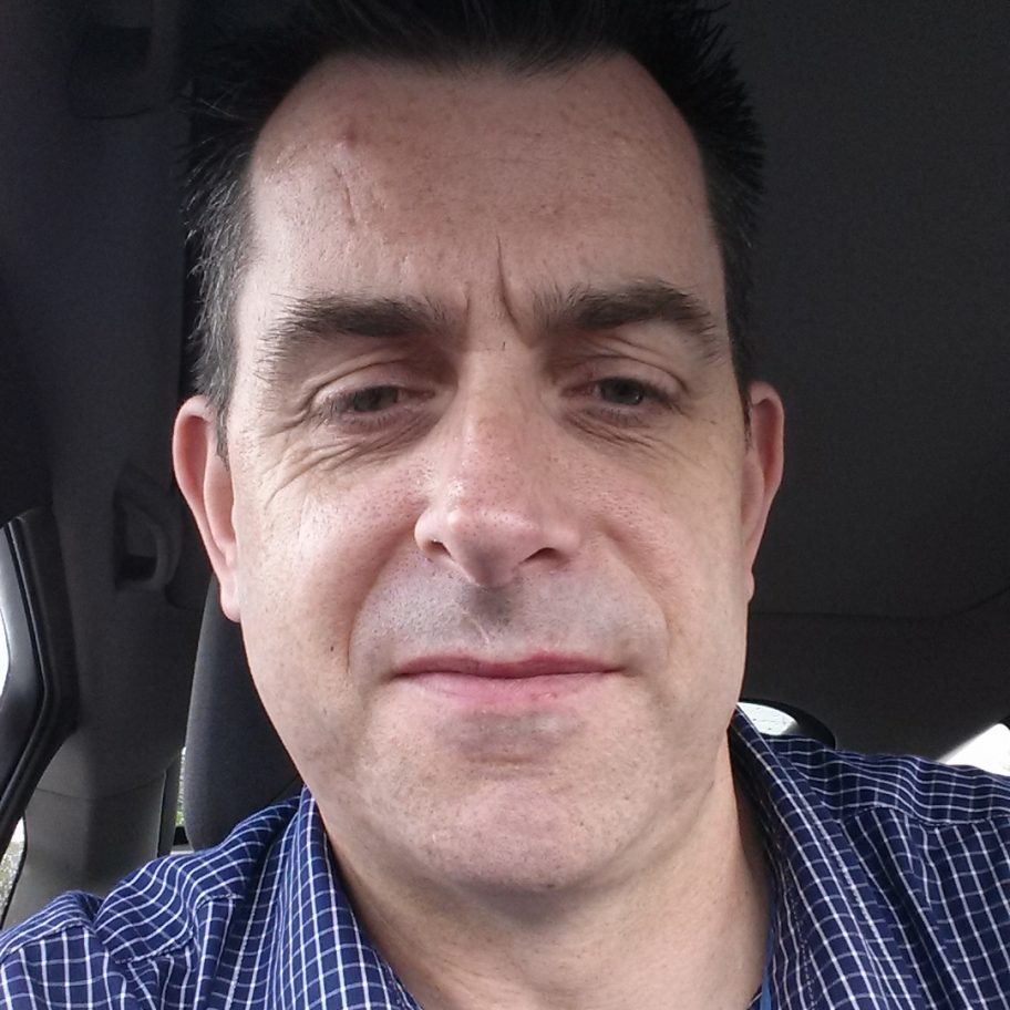 A middle-aged man with short dark hair and wearing a blue and white checkered shirt is taking a selfie inside a car. He has a neutral expression on his face. The car's interior is partially visible in the background.