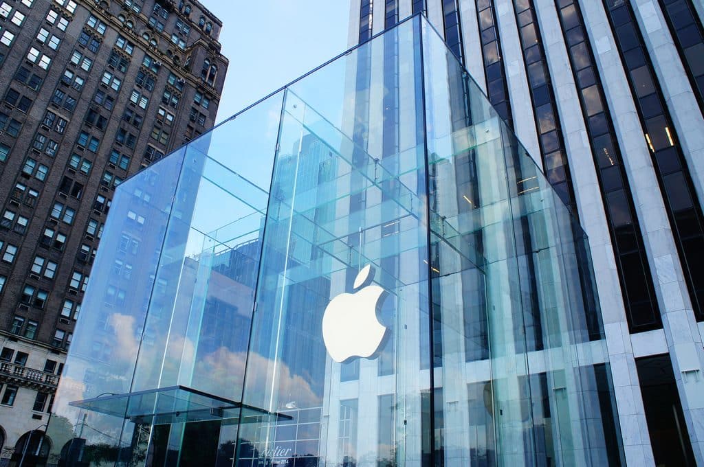 A large glass cube structure with a glowing white Apple logo in its center stands in front of tall buildings, showcasing Apple's ambitious design. The cube is the entrance to an Apple Store, with a staircase leading down into it. The sky is clear, reflecting off the glass walls.