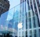 A large glass cube structure with a glowing white Apple logo in its center stands in front of tall buildings, showcasing Apple's ambitious design. The cube is the entrance to an Apple Store, with a staircase leading down into it. The sky is clear, reflecting off the glass walls.