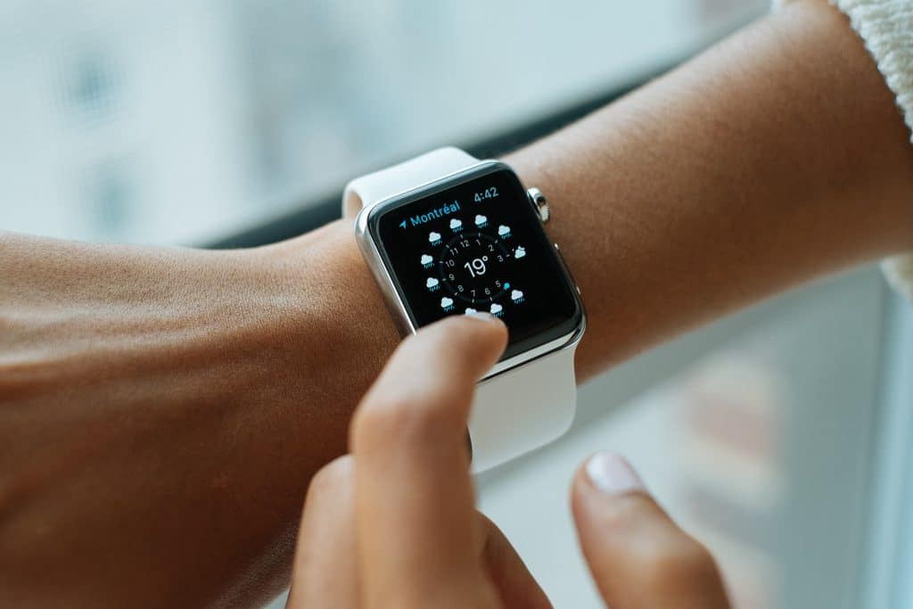 Close-up of a person interacting with an Apple Watch Series 2 on their wrist. The smartwatch display shows a weather application with current conditions for Montreal, highlighting a temperature of 19 degrees. The smartwatch, equipped with new batteries, has a white band.