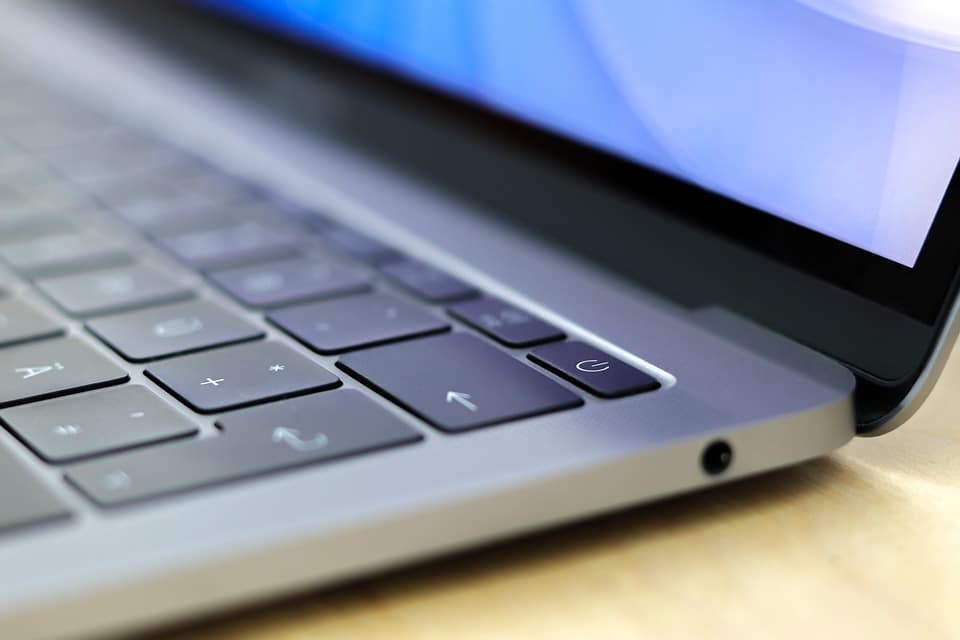 Close-up of a sleek, modern Mac keyboard and part of its screen. The focus is on the keys, with the power button particularly prominent. The laptop has a metallic finish and is placed on a light-colored surface. The screen displays a blue-toned background, emphasizing Apple’s signature design.