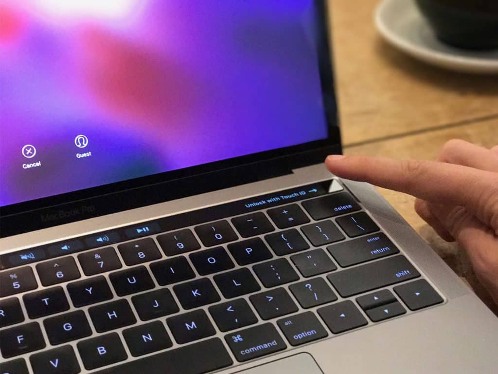 A person's finger is touching the fingerprint sensor on a 2018 MacBook Pro with the login screen displayed. The keyboard and Touch Bar are visible, and there is a blurred background with a cup partially in view, offering a glimpse of what you can expect from this sleek device.