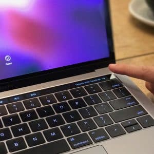 A person's finger is touching the fingerprint sensor on a 2018 MacBook Pro with the login screen displayed. The keyboard and Touch Bar are visible, and there is a blurred background with a cup partially in view, offering a glimpse of what you can expect from this sleek device.