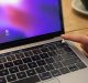 A person's finger is touching the fingerprint sensor on a 2018 MacBook Pro with the login screen displayed. The keyboard and Touch Bar are visible, and there is a blurred background with a cup partially in view, offering a glimpse of what you can expect from this sleek device.