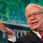 An elderly man with glasses, wearing a suit and tie, gestures with his right hand while speaking at an event. A green and blue backdrop with various symbols and text is visible behind him, reminiscent of Warren Buffett discussing Apple's $1 trillion valuation.