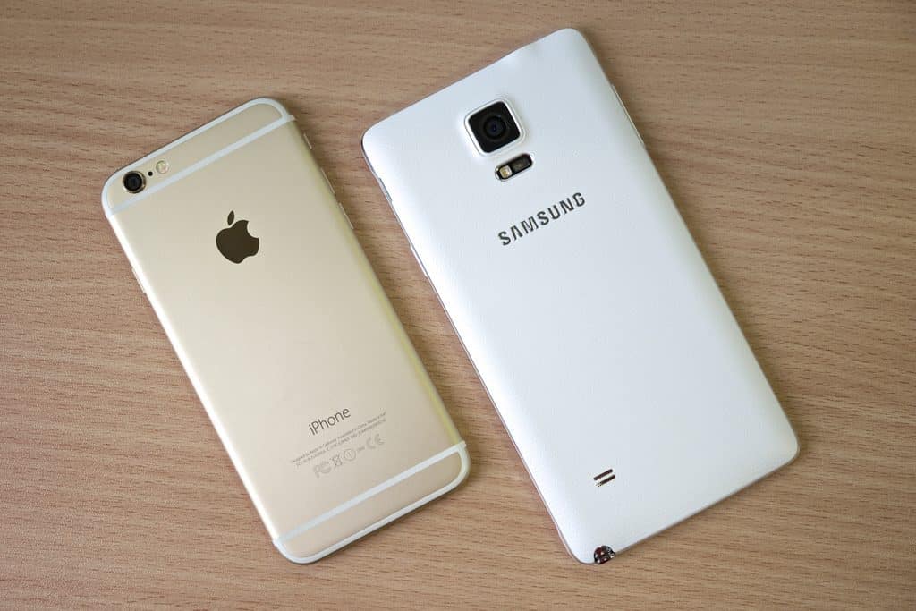 A gold Apple iPhone with the logo displayed on the back is placed next to a white Samsung smartphone, its logo visible. Both phones are resting on a wooden surface. The iPhone has a smooth metallic finish, while the Samsung phone features a textured back, like two contenders awaiting a jury decision.