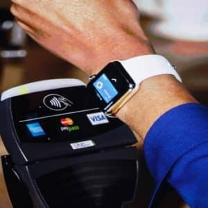 A person wearing a smartwatch with a white band is making an Apple Pay contactless payment at a terminal. The terminal displays logos for MasterCard, paypass, Visa, and other payment options. A coffee cup is visible on the right side of the image.