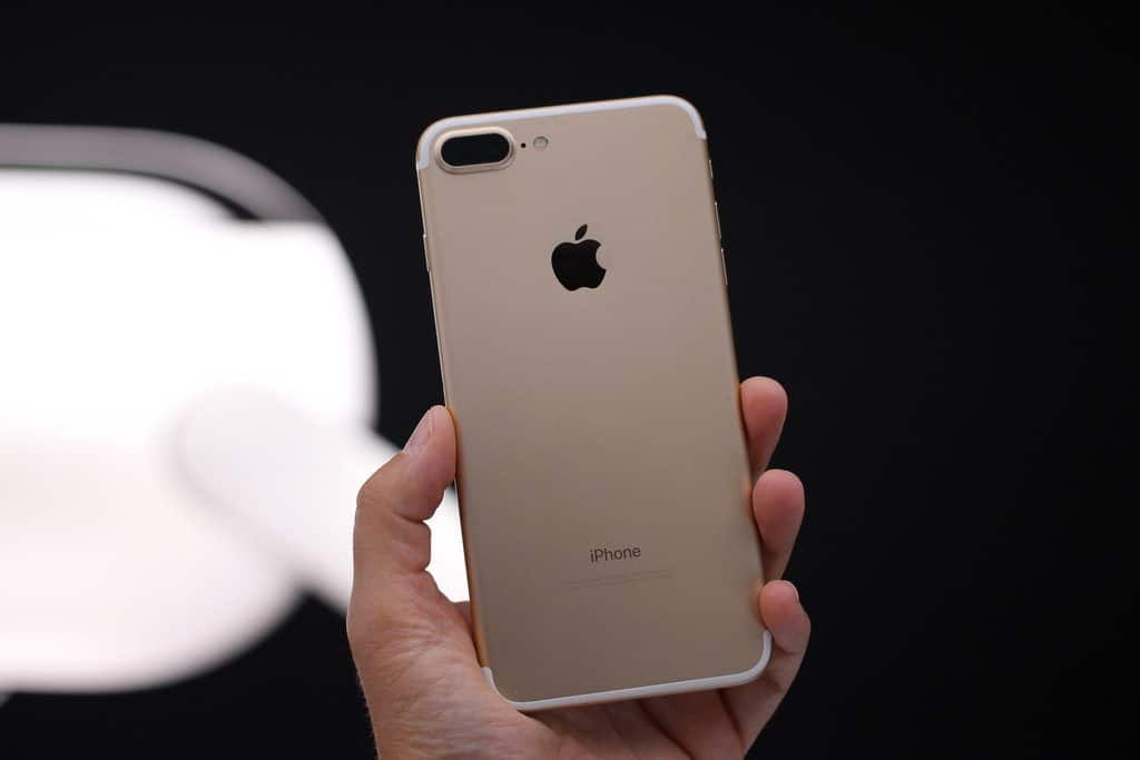 A hand is holding a gold-colored iPhone 7 Plus, showing the back of the device. The Apple logo is centered near the top, and the smartphone features a dual-camera system in the upper left corner. The background is blurred, highlighting the focus on consumer satisfaction with the iPhone 7 Plus.