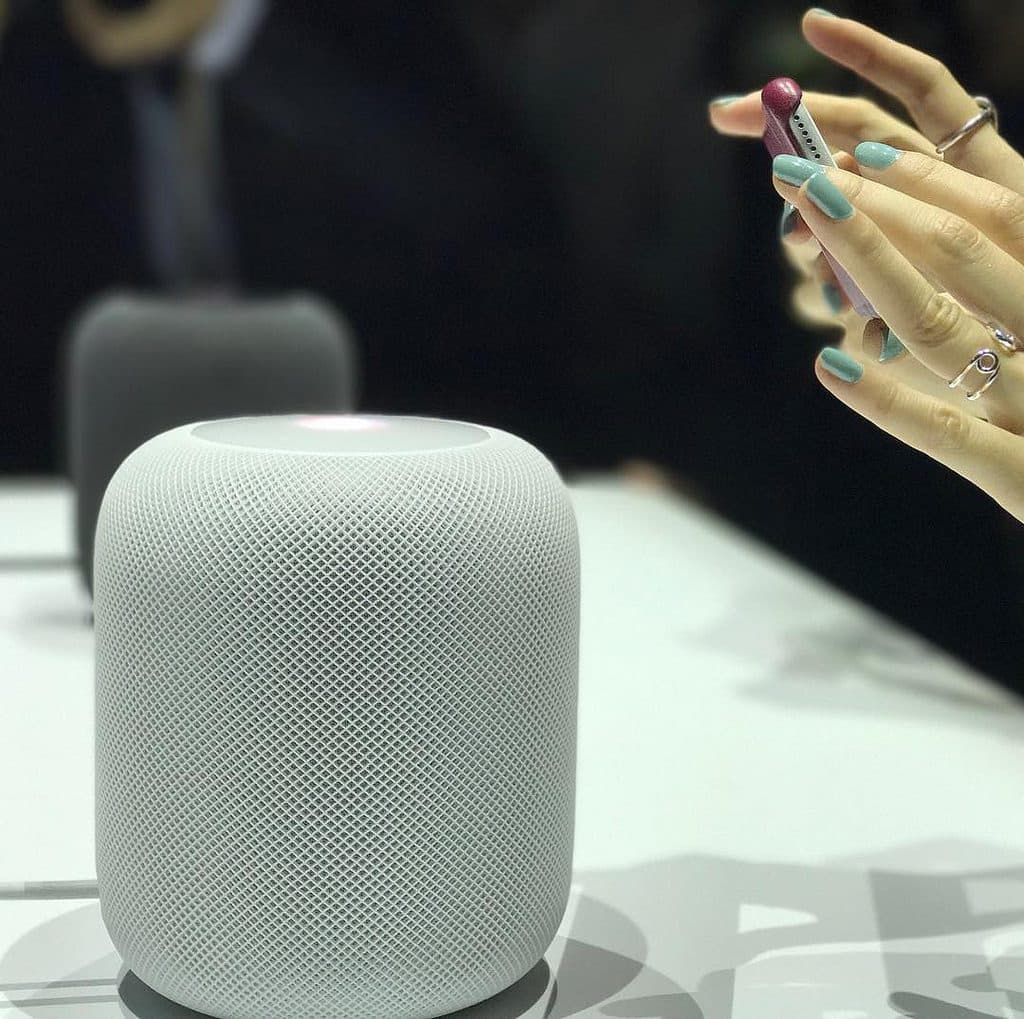 A white, cylindrical smart speaker is placed on a table. To the right, a person's hands with mint green nails are holding a small object with a red tip. Another similar speaker is blurred in the background, reminiscent of the gadgets showcased at WWDC 2018.