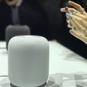 A white, cylindrical smart speaker is placed on a table. To the right, a person's hands with mint green nails are holding a small object with a red tip. Another similar speaker is blurred in the background, reminiscent of the gadgets showcased at WWDC 2018.