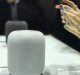 A white, cylindrical smart speaker is placed on a table. To the right, a person's hands with mint green nails are holding a small object with a red tip. Another similar speaker is blurred in the background, reminiscent of the gadgets showcased at WWDC 2018.