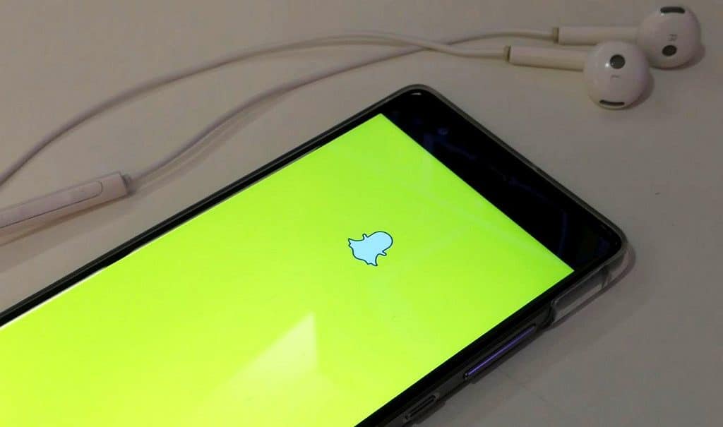 A smartphone screen displays the Snapchat logo on a yellow background, emphasizing its privacy features. Next to the phone, a pair of white earphones with an in-line remote are laid out on a white surface.