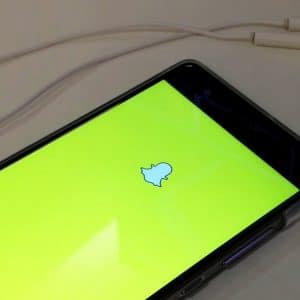 A smartphone screen displays the Snapchat logo on a yellow background, emphasizing its privacy features. Next to the phone, a pair of white earphones with an in-line remote are laid out on a white surface.