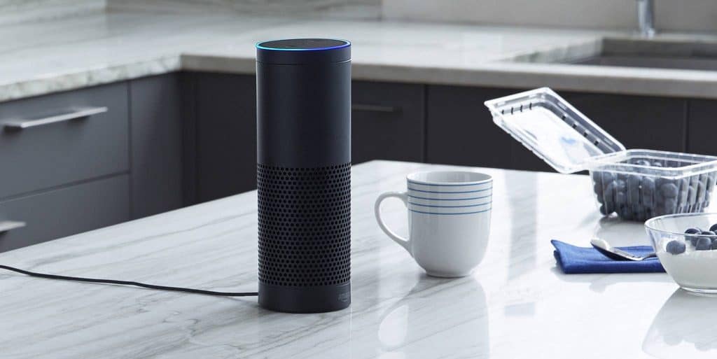 A smart speaker, featuring Amazon Alexa, is placed on a marble kitchen countertop next to a white mug with blue stripes, a clear plastic container of blueberries, and a blue cloth napkin. In the background are dark kitchen cabinets and part of a stainless steel sink.