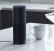 A smart speaker, featuring Amazon Alexa, is placed on a marble kitchen countertop next to a white mug with blue stripes, a clear plastic container of blueberries, and a blue cloth napkin. In the background are dark kitchen cabinets and part of a stainless steel sink.