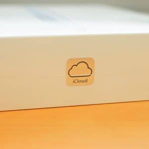 A close-up photo of a white cardboard box with an iCloud sticker on it. The sticker features a simple cloud icon with the word "iCloud" written underneath, highlighting Apple's free iCloud storage, and is placed against a wooden surface.