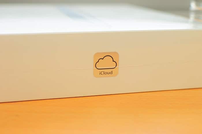 A close-up photo of a white cardboard box with an iCloud sticker on it. The sticker features a simple cloud icon with the word "iCloud" written underneath, highlighting Apple's free iCloud storage, and is placed against a wooden surface.