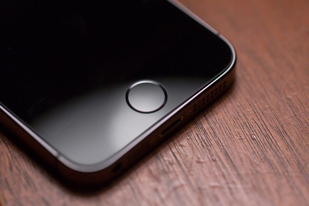 A close-up of the lower portion of an iPhone SE2 placed on a wooden surface. The image features the device's home button and part of the front screen. The black screen contrasts with the brown wood texture, exceeding expectations in design.