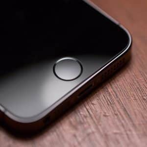 A close-up of the lower portion of an iPhone SE2 placed on a wooden surface. The image features the device's home button and part of the front screen. The black screen contrasts with the brown wood texture, exceeding expectations in design.