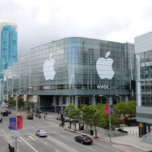 A large glass-fronted convention center adorned with Apple's logo prepares to host the WWDC 2011 event. Numerous people are walking along the adjacent streets, and a tall modern building is visible in the background under a cloudy sky, reminiscent of anticipation felt before an Apple keynote.