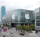 A large glass-fronted convention center adorned with Apple's logo prepares to host the WWDC 2011 event. Numerous people are walking along the adjacent streets, and a tall modern building is visible in the background under a cloudy sky, reminiscent of anticipation felt before an Apple keynote.