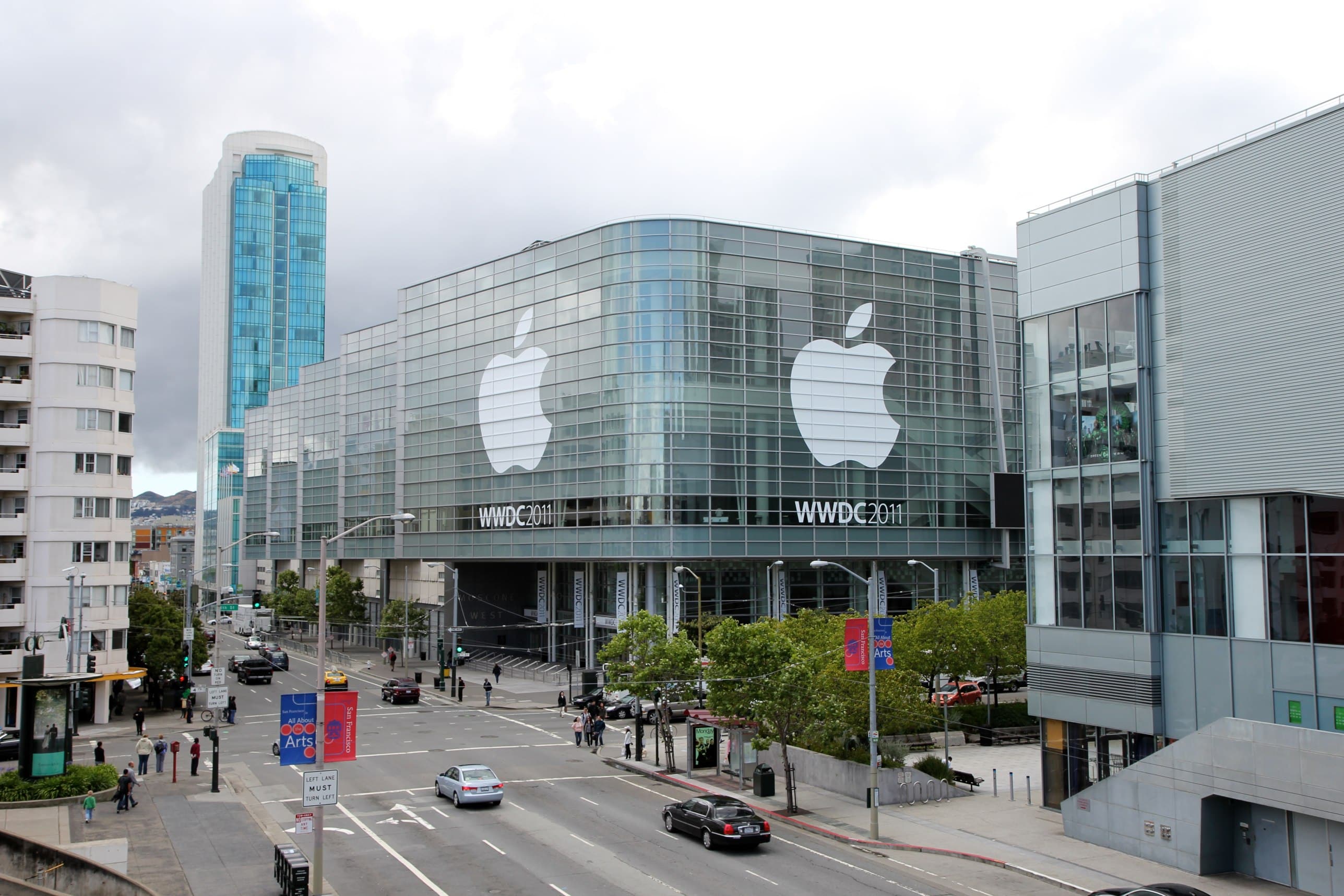 A large glass-fronted convention center adorned with Apple's logo prepares to host the WWDC 2011 event. Numerous people are walking along the adjacent streets, and a tall modern building is visible in the background under a cloudy sky, reminiscent of anticipation felt before an Apple keynote.