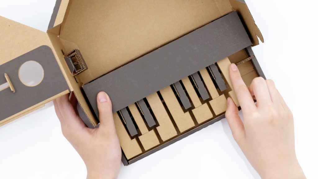 A child's hands are playing with a Nintendo Labo model of a piano. Constructed from brown and black cardboard, it features black keys and a minimalist design. The right hand presses a key while the left holds the creation steady.