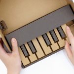 A child's hands are playing with a Nintendo Labo model of a piano. Constructed from brown and black cardboard, it features black keys and a minimalist design. The right hand presses a key while the left holds the creation steady.