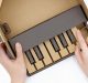 A child's hands are playing with a Nintendo Labo model of a piano. Constructed from brown and black cardboard, it features black keys and a minimalist design. The right hand presses a key while the left holds the creation steady.