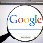 A magnifying glass focuses on the Google logo displayed on an iPhone screen. The search bar and 'Google-Suche' button are visible below, along with part of the Chrome browser window and tabs above, subtly emphasizing the blend of technology and privacy.