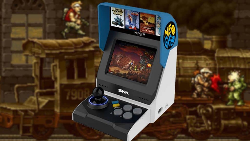 A SNK Mini Neo Geo arcade machine with an SNK logo on the front features a joystick, buttons, and is displaying an action game on its screen. The backdrop is an in-game scene with two characters on a moving train. Box art for various games is visible on top of the machine.