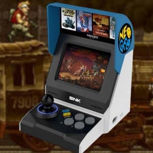 A SNK Mini Neo Geo arcade machine with an SNK logo on the front features a joystick, buttons, and is displaying an action game on its screen. The backdrop is an in-game scene with two characters on a moving train. Box art for various games is visible on top of the machine.