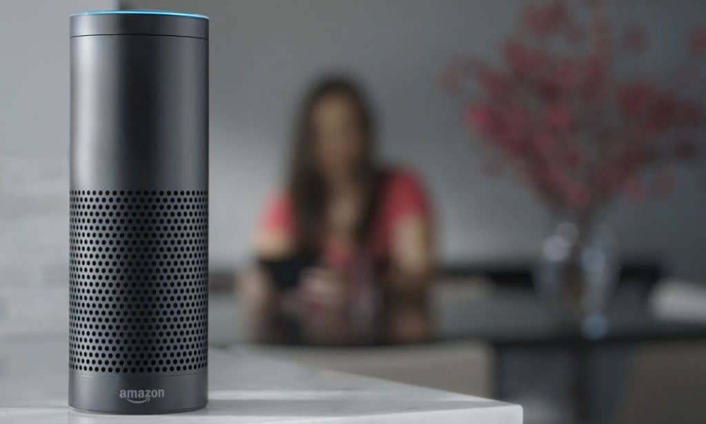 A black Amazon Echo smart speaker with Alexa sits on a white counter in focus. In the blurred background, a person wearing a red shirt is seated at a table with a vase of flowers.