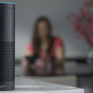 A black Amazon Echo smart speaker with Alexa sits on a white counter in focus. In the blurred background, a person wearing a red shirt is seated at a table with a vase of flowers.