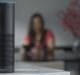 A black Amazon Echo smart speaker with Alexa sits on a white counter in focus. In the blurred background, a person wearing a red shirt is seated at a table with a vase of flowers.