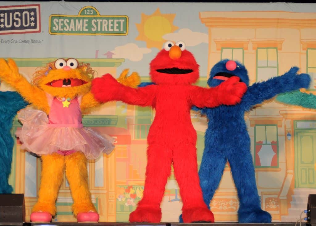 Three Sesame Street characters stand together with their arms open wide. From left to right, they are a yellow character wearing a pink dress, a red character, and a blue character. The backdrop resembles a colorful Sesame Street neighborhood, showcasing the charm of kids' programming by Sesame Workshop.