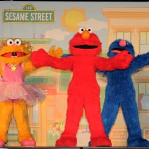 Three Sesame Street characters stand together with their arms open wide. From left to right, they are a yellow character wearing a pink dress, a red character, and a blue character. The backdrop resembles a colorful Sesame Street neighborhood, showcasing the charm of kids' programming by Sesame Workshop.